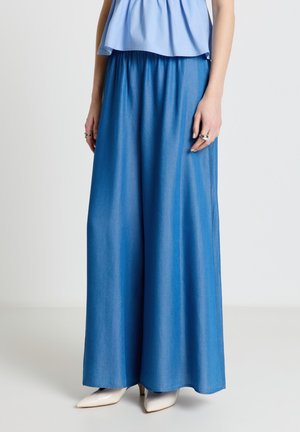 Donna che indossa pantaloni in denim blu a gamba larga abbinati a tacchi bianchi a punta e top azzurro senza maniche.