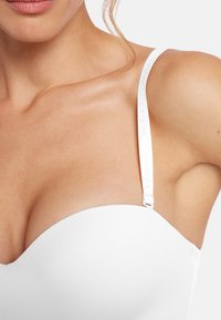 Vit axelbandslös bh med slät tyg, en formad siluett och justerbara band där varumärket diskret visas.