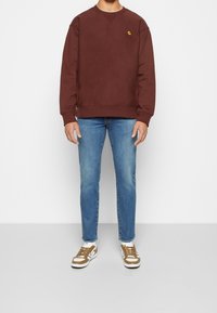 Brun sweatshirt med rund hals och ribbade ärmar, parat med blå jeans och bruna sneakers med vita accenter. Enkel design.