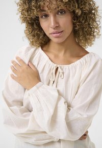 Light cream blouse with textured fabric, elasticated neckline, tie detail, and voluminous sleeves. Subtle sheen adds visual interest.