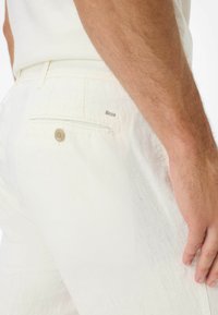 Pantaloni bianchi di lino con una tasca posteriore con chiusura a bottone e un piccolo cartellino in metallo. Il tessuto presenta una finitura leggermente strutturata.