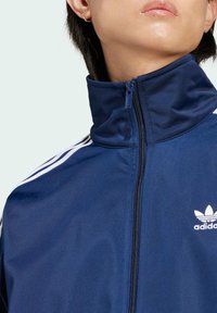 Veste de survêtement Adidas bleu marine avec col montant, fermeture éclair intégrale et accents blancs en trois bandes sur les manches. Tissu texturé avec logo.
