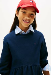 Felpa a coste blu navy con design peplum e un piccolo logo; indossata sopra una camicia a righe azzurre con dettagli ai bottoni. Cappellino rosso indossato sulla testa.