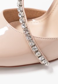 Beige lackläder högklackad sko med en tunn ankelsnodd prydd med klara rhinestones. Rund tå och slät yta.