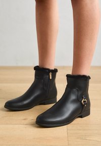 Bottines noires en cuir lisse, avec un revers en fourrure, fermeture éclair latérale et petit accent en métal doré. Semelle plate avec peu de relief.