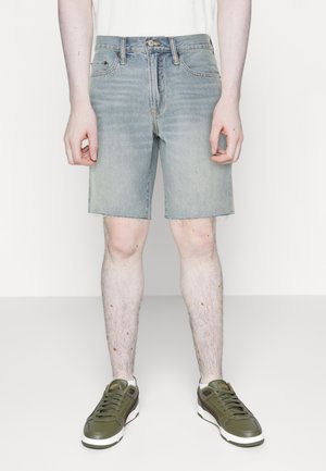 Short en denim bleu clair avec un ourlet effiloché, conception à cinq poches et fermeture à bouton. Porté avec des baskets vert olive.
