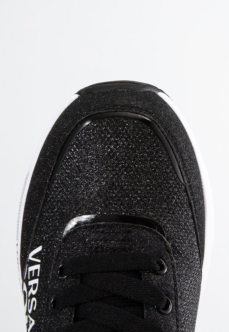 Zapatilla deportiva negra con parte superior texturizada, detalles en negro brillante y suela blanca. Presenta cordones negros y detalle del logo en el lateral.