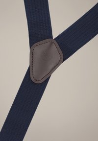 Granatowe elastyczne szelki z teksturowanym wzorem oraz brązową skórzaną naszywką na łączeniu, na której znajduje się tłoczony logo.