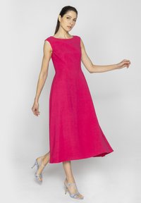 Fuksjowa, bezrękawnikowa sukienka z dopasowaną górą i rozkloszowanym dołem, wykonana z lekkiego materiału. Zestawiona z jasnoniebieskimi sandałami na obcasie.
