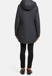 Personne portant un manteau parka à capuche gris foncé, un pantalon slim noir et des baskets noires, se tenant de dos sur un fond clair.