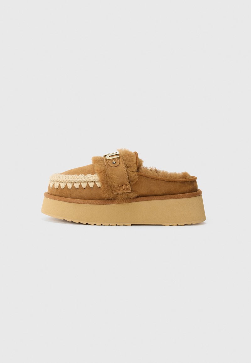 Brown suede slip-on shoes with fur lining, decorative woven trim, gold buckle accent, and a thick, textured sole.