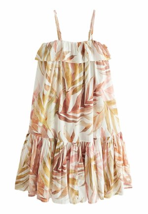 Nainen, joka pukee hihattoman, laskeutuvan midi-mekon, jossa on ruskeita ja beigejä lehtikuvioita, ja ruskeat tasaiset sandaalit, seisoo minimalististen vaaleiden seinien edessä.