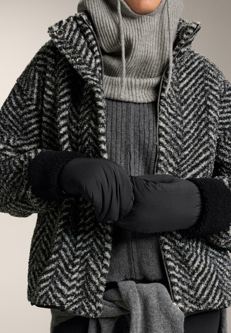 Jachetă cu un model herringbone în nuanțe de negru și gri, cu guler înalt, asortată cu un pulover gri cu dungi și mănuși negre căptușite. Materiale texturate observate.