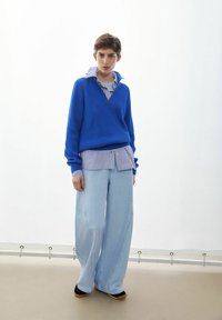 Blauer gestrickter Pullover mit Reißverschluss am Hals, darüber getragen ein gestreiftes Hemd, kombiniert mit weit geschnittenen, hellen Jeans und dunklem Schuhwerk.