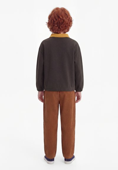 Un enfant aux cheveux roux bouclés est de dos, portant un haut à manches longues foncé avec un col moutarde, un pantalon en velours côtelé marron et des chaussures violettes.