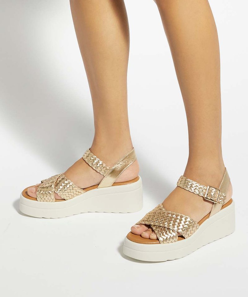 Jambes portant des sandales à plateforme avec brides tressées métalliques dorées et semelles blanches épaisses sur un fond clair uni.