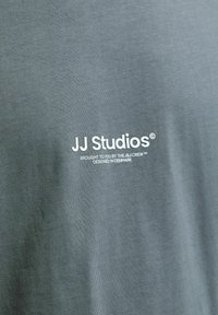 Graues Baumwoll-T-Shirt mit einem weißen aufgedruckten Logo "JJ Studios" und dem Text "Brought to you by the J.J. Crew™ Designed in Denmark."