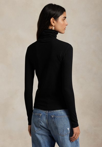 Naine, kes kannab keha järgivat musta kaelusega kampsunit ja siniseid teksapükse, seisab seljaga kaamera poole tühja tausta ees.