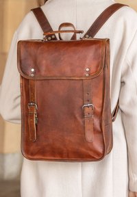 Mochila de cuero marrón con un diseño estructurado, herrajes plateados, correas ajustables y un asa en la parte superior. Presenta detalles de costura en los bordes.