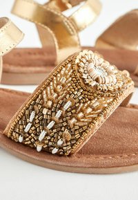 Goldene Ledersandalen mit einem dekorativen, mit Perlen besetzten Riemen und komplizierten Mustern. Wildlederfußbett mit weicher Textur und brauner Nuance.
