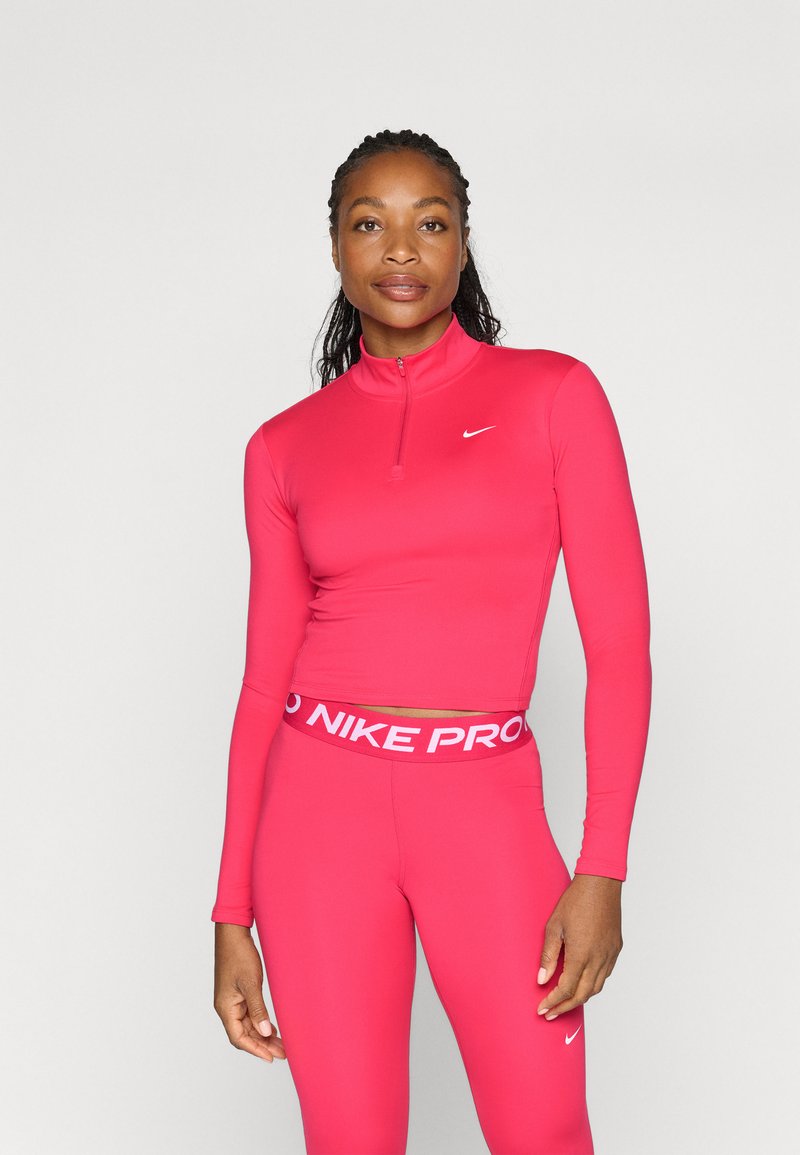 Feszített rózsaszín sportfelső, félig cipzárral és hosszúujjakkal, amelyet hozzá illő, magas derekú leggings-szel párosítanak, fehér márkázott derékpánttal.