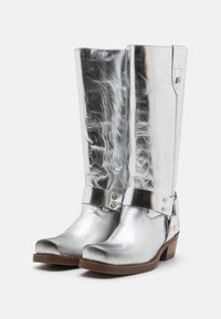 Bottes argentées métalliques montantes avec une finition en cuir lisse, un bout arrondi et un accent en boucle en métal à la cheville. Le talon en bois ajoute de la stabilité.