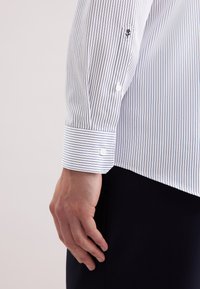 Camicia bianca a strisce verticali blu. Presenta polsini con bottoni e un logo discreto sulla manica. Il materiale appare liscio e fresco.