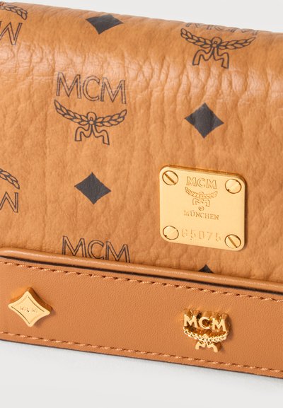 Section de sac MCM en cuir fauve avec motif de logo noir, plaque métallique dorée gravée MCM München et numéro de série, et clous MCM dorés.