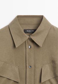Khaki cotton shirt with a buttoned collar, chest pockets, and silver hardware. Features a textured, solid fabric with contrasting stitching.