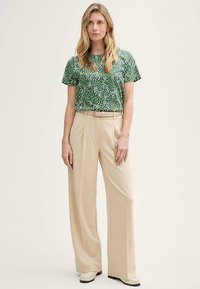 Haut vert à manches courtes avec des points blancs éparpillés, associé à un pantalon beige taille haute à jambes larges et une ceinture assortie. Porté avec des chaussures claires.