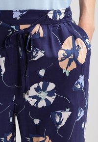 Námořnické modré květinové kalhoty s elastickým pasem, detaily na šňůrku a velkými světlými květinovými vzory. Hladká textura látky.