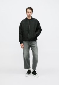 Μαύρο μπουφάν bomber με καπιτονέ σχέδιο και φερμουάρ, συνδυασμένο με ανοιχτό γκρι τζιν με ίσια γραμμή και μαύρα αθλητικά παπούτσια με λευκές σόλες.