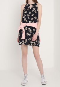 Czarna sukienka w kwiaty z dekoltem halter, wycięciem w kształcie łezki i rozkloszowanym dołem, do której dobrano różowy sweter związany w pasie oraz białe sportowe buty wysokie.