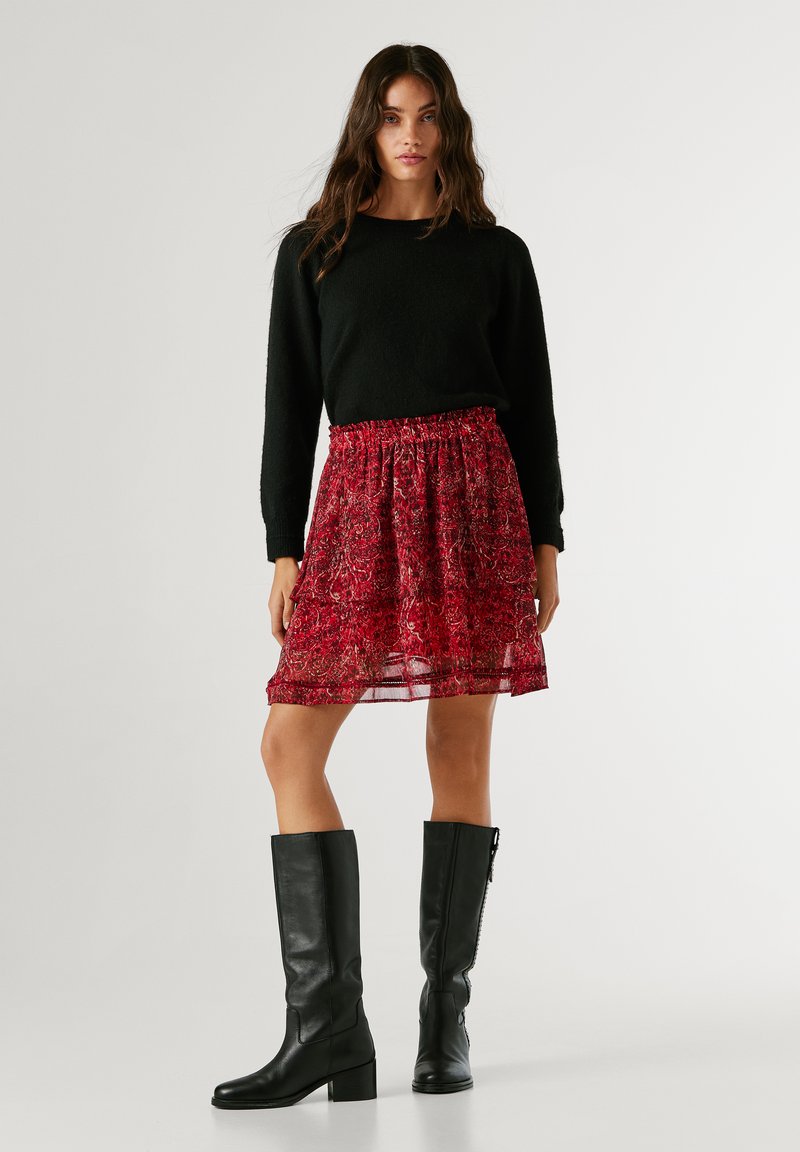 Musta villapaita, punaisella kukkakuosisella hameella, jossa on joustava vyötärö, läpinäkyvä päällyskerros ja polven korkuiset mustat nahkasaappaat, joissa on paksut korot.