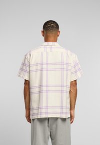 Camicia a maniche corte di colore crema con un motivo a quadretti lilla, caratterizzata da un tessuto testurizzato e orlo dritto. Vista posteriore, colletto visibile.