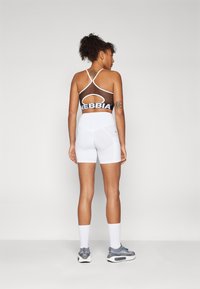 Pantaloni sportivi bianchi con vita alta e reggiseno sportivo in rete con una fascia nera. La modella indossa scarpe da ginnastica grigie e calzini bianchi.