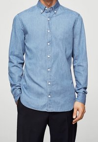 Hellblaues Denimhemd mit langen Ärmeln, Knopfleiste am Kragen und weißen Knöpfen. Figurbetonter Schnitt mit abgerundetem Saum und glatter Textur.