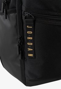 Rucsac din material textil negru, cu un exterior texturat, branduire aurie, fermoare netede și un tag proeminent cu logo-ul Jordan.