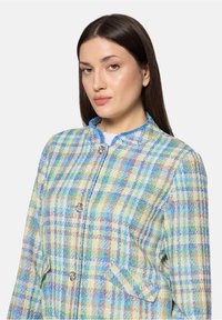 Kurtka w wielokolorową kratę z teksturowanej tkaniny, z niebieskimi akcentami, okrągłym dekoldem i dwoma przednimi kieszeniami, zapinana na metalowe guziki.