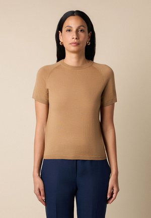 Kobieta o długich czarnych włosach, ubrana w beżowy krótki sweter z dzianiny, złote kolczyki-koła i ciemnoniebieskie spodnie, na jednolitym tle.