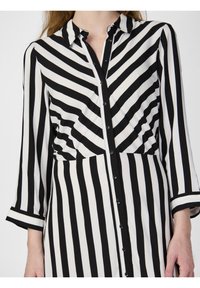 Femme portant une robe chemise rayée noire et blanche, à rayures verticales et diagonales, manches trois-quarts et col.