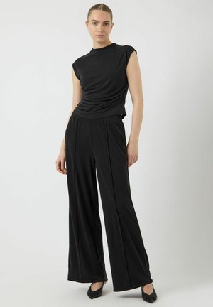 Donna in piedi con un top nero senza maniche con lato arricciato e pantaloni neri a gamba larga abbinati a scarpe nere a punta, su uno sfondo bianco.