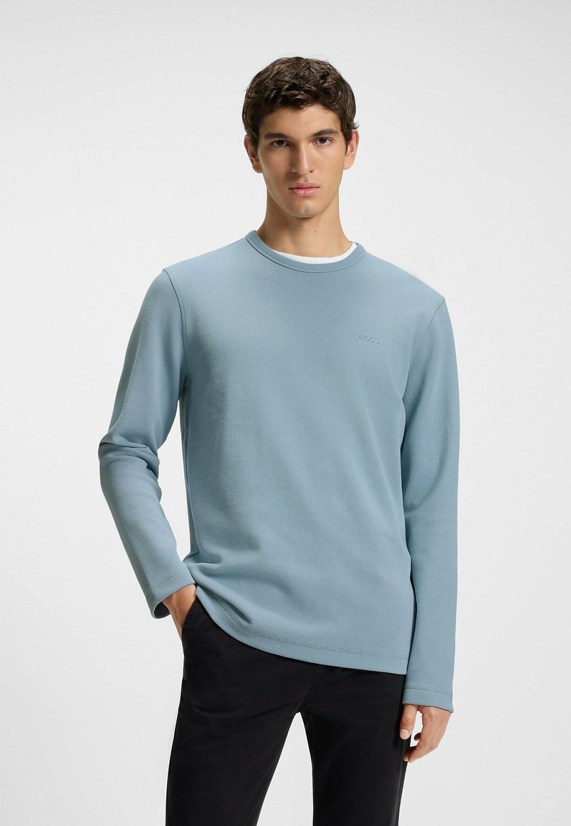 Light blue long-sleeve shirt with a textured pattern and round neck. Worn over black pants, featuring subtle branding on the front.