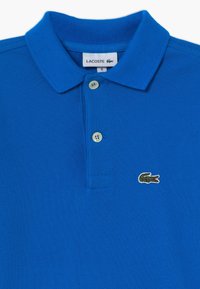 Blå polo skjorta i texturerad tyg, med klassisk krage och tre knappar. Lacoste-logotyp broderad i grönt med ett rött inslag.