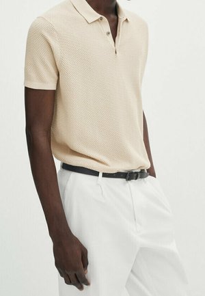 Beige textured polo shirt with short sleeves, two-button placket, worn with white trousers and a black belt. Simple design, lightweight fabric.