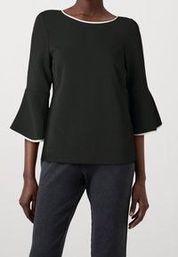 Černý top s lodičkovým výstřihem, tříčtvrtečními zvonovými rukávy a bílými páskami. Vytvořený v kombinaci s texturovanými tmavými kalhotami s proužky.