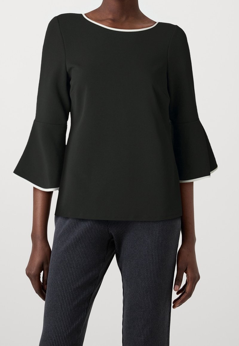 Černý top s lodičkovým výstřihem, tříčtvrtečními zvonovými rukávy a bílými páskami. Vytvořený v kombinaci s texturovanými tmavými kalhotami s proužky.