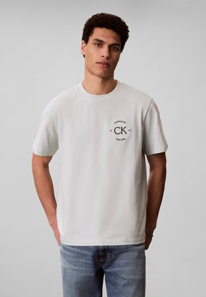 Mężczyzna ubrany w białą koszulkę Calvin Klein z logo na piersi i niebieskie dżinsy, stojący na tle jednolitego szarego tła.