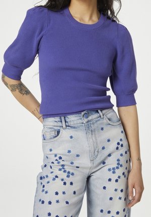 Woman wearing a purple ribbed short-sleeve sweater with shoulder buttons and light-wash jeans featuring blue floral prints.