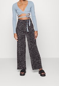 Jasnoniebieska bluzka z długim rękawem, zapinana na zakładkę z detalem w postaci wiązania, oraz czarne spodnie w szeroką nogawkę z drobnym białym wzorem kwiatowym. Czarne buty na platformie.